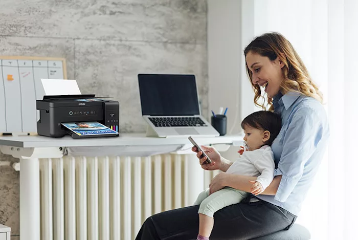 Mulher com bebé ao colo e secretária com computador portátil e impressora ecotank em segundo plano