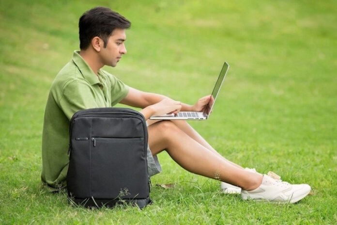 Rapaz sentado na relva com um computador portátil e mochila de portátil