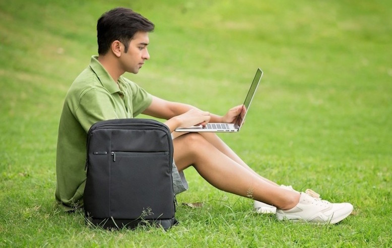 Rapaz sentado na relva com um computador portátil e mochila de portátil